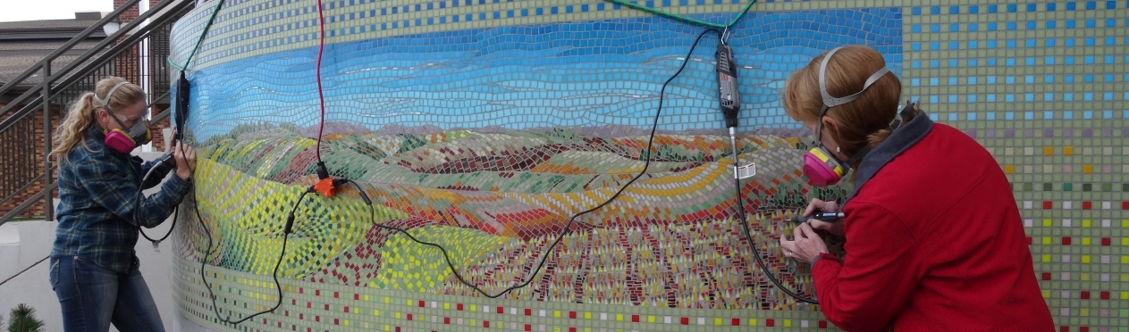 Two women work on installing a mosaic artwork that is a landscape of Eastern Washington.