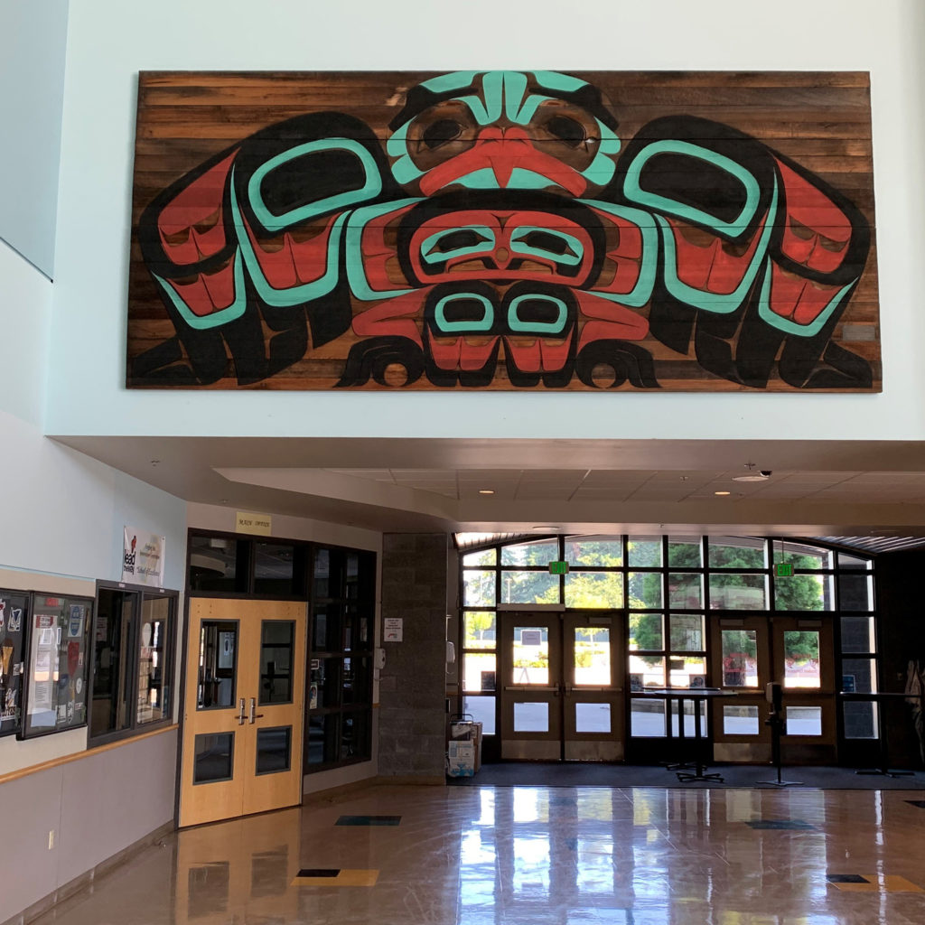 A double-height room with a large carved and painted wooden mural placed above the entry-way. The mural depicts a bird form in coast salish style formline design.