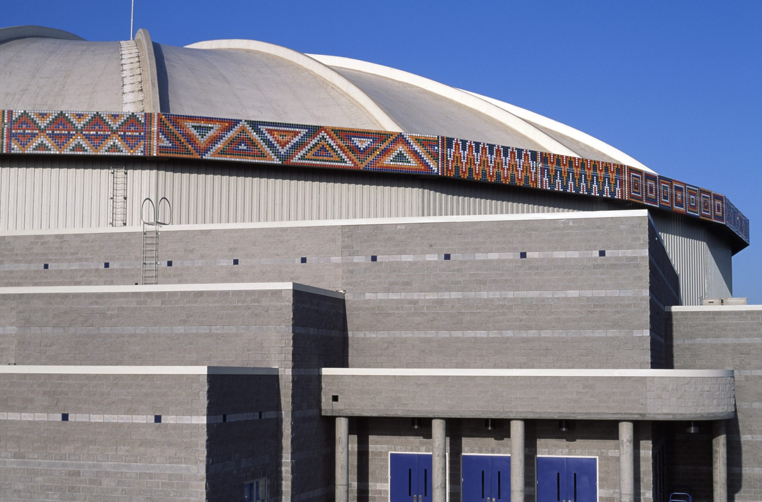 Partial view of a band of geometric patterns made of thousands of 3-inch industrial highway reflectors that encircle the roof of a large concrete building. There is a very blue sky behind the SunDome building.