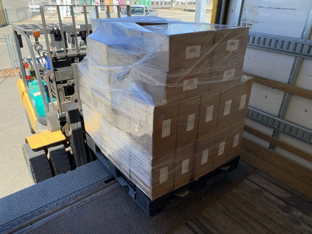 A forklift removes a shrik-wrapped palette of cardboard boxes from the back of a truck. The boxes contain the reflectors for the restorations.